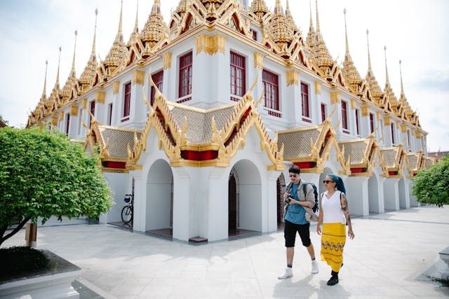 Dos turistas caminan junto a un templo con detalles dorados en Bangkok, Tailandia. Título: Explorando la majestuosidad de los templos tailandeses.