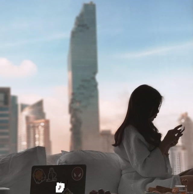Mujer con bata blanca usando su teléfono en una habitación con vista al skyline de Bangkok, con el rascacielos MahaNakhon de fondo.