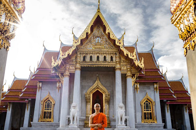 Monje budista con túnica naranja posando frente al Templo del Mármol en Bangkok, Tailandia, destacando la arquitectura tradicional tailandesa.