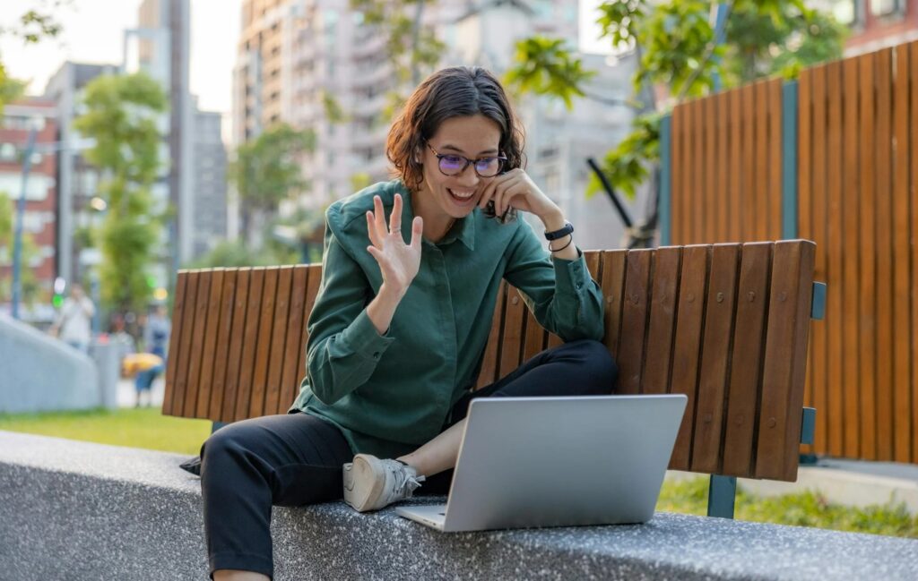 Persona sonriente realizando una videollamada desde un espacio al aire libre con su ordenador portátil, representando la flexibilidad del teletrabajo.