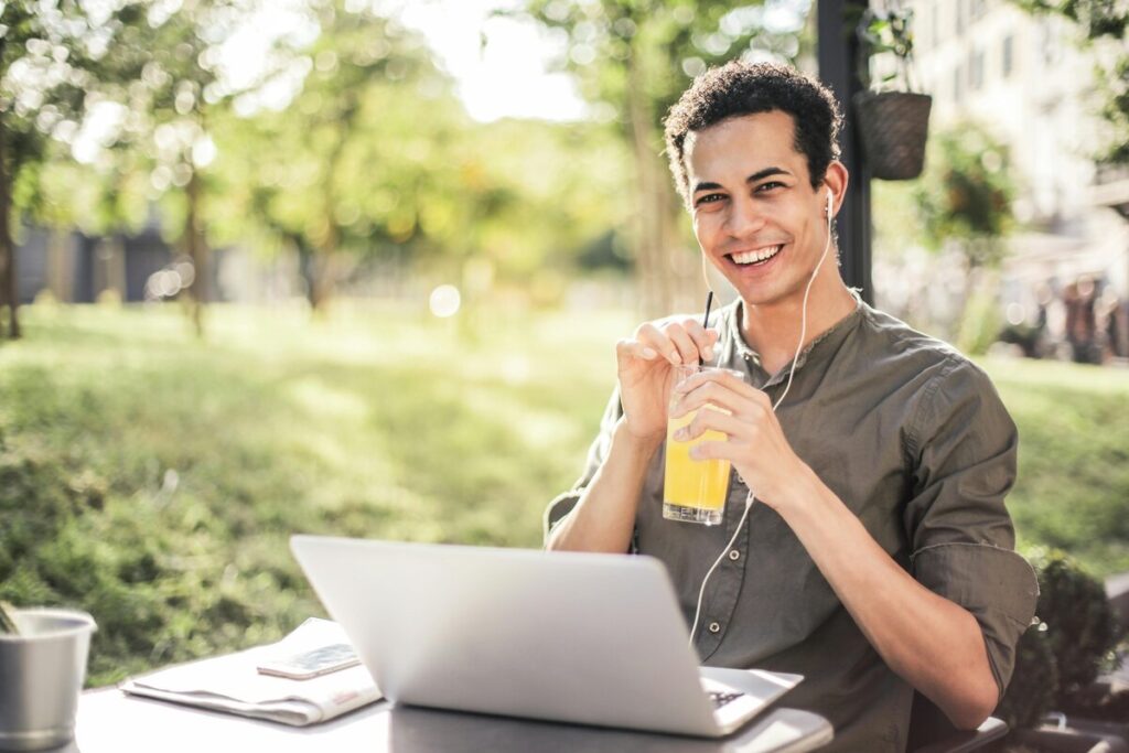 Hombre sonriendo trabajando en un portátil al aire libre, con auriculares y sosteniendo un vaso de jugo en un entorno verde y soleado.