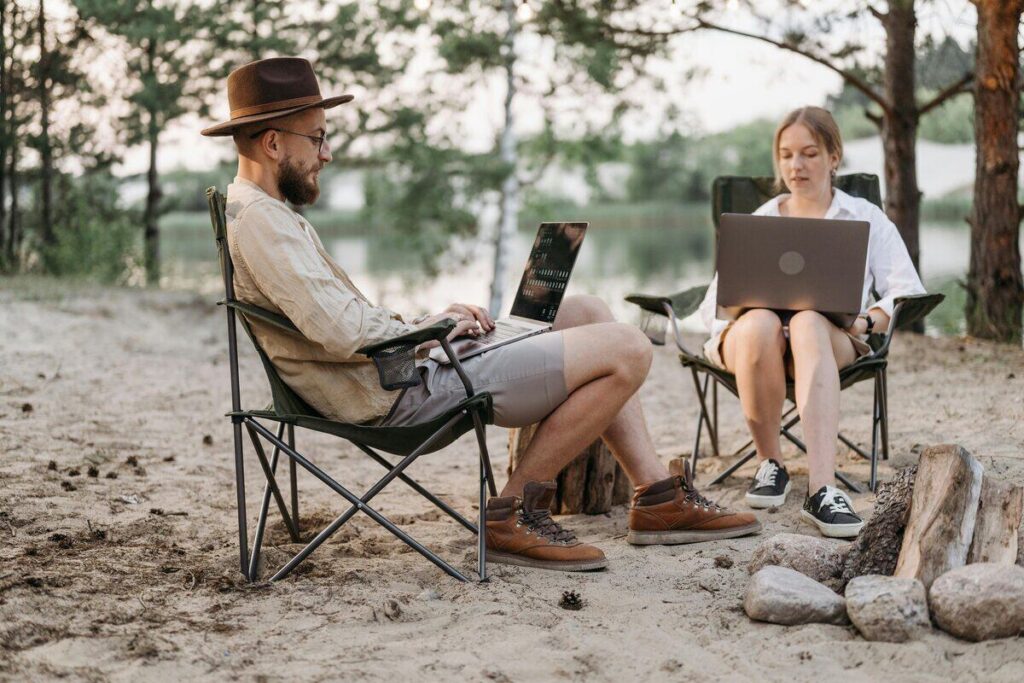 Dos personas trabajando en remoto con sus ordenadores portátiles en un entorno natural, sentados en sillas plegables junto a una fogata en un bosque junto al agua.