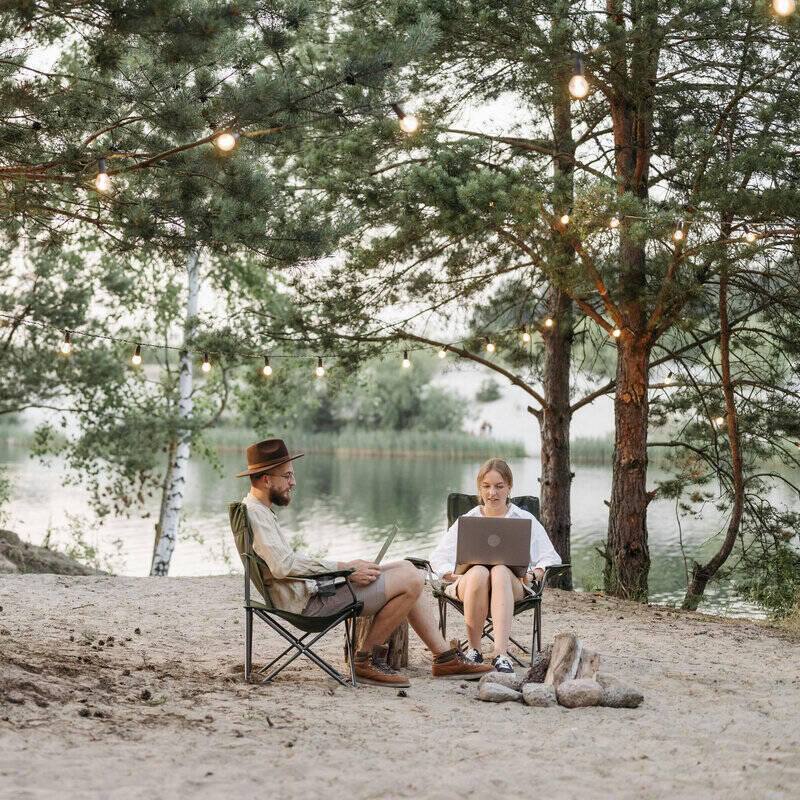 Pareja teletrabajando al aire libre junto a un lago, rodeados de árboles y luces decorativas, disfrutando de la flexibilidad del trabajo remoto en plena naturaleza.