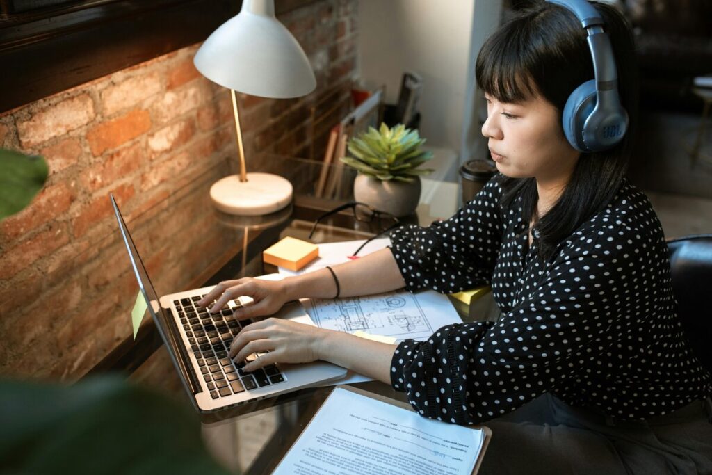 Mujer teletrabajando desde un espacio acogedor, con auriculares y portátil, rodeada de documentos, una planta y una lámpara de escritorio.